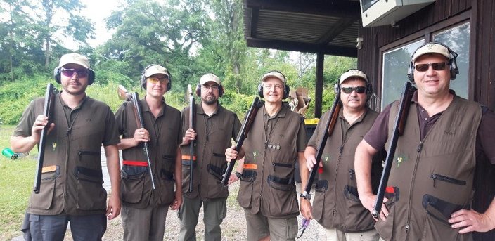 Sechs Personen stehen mit Gewehren auf einem Schießstand, tragen Schutzbrillen und Gehörschutz. Sie sind in Uniformen gekleidet, in einer bewaldeten Umgebung mit einem Holzgebäude im Hintergrund.